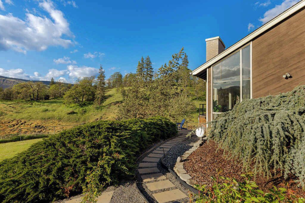 Une maison moderne avec un chemin de pierres menant à une porte en bois, entourée d’une végétation luxuriante et d’arbres.