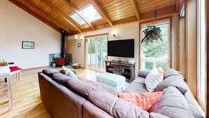 A living room with a wooden ceiling, a large window, a television, and a sofa. - Tree-lined coastal home with wood fireplace, yard, W&D, & games - dog-friendly (Manzanita)