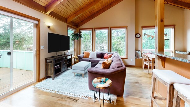 A spacious living area with a high ceiling, a large window, a sofa, a TV, and a dining area. - Tree-lined coastal home with wood fireplace, yard, W&D, & games - dog-friendly (Manzanita)