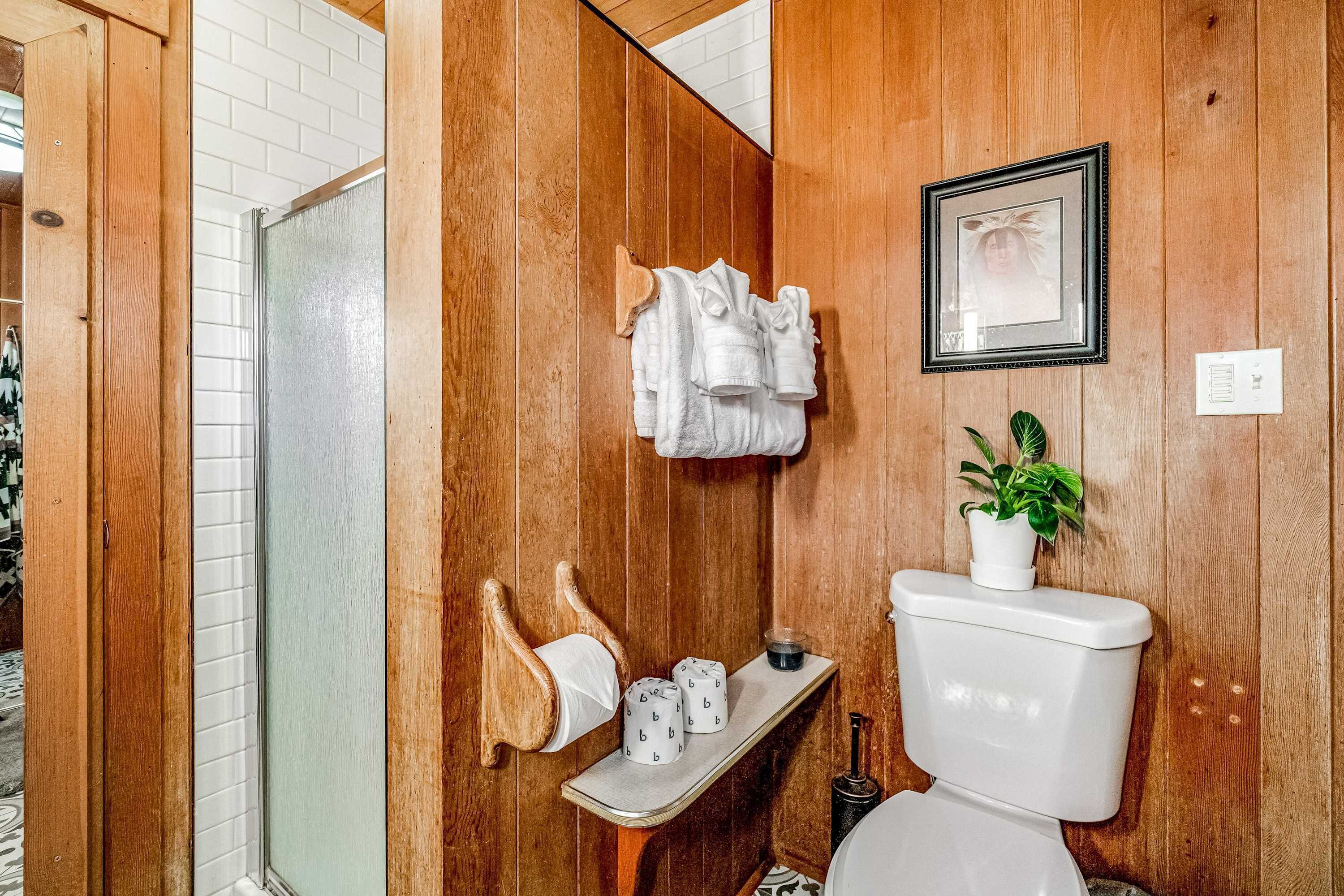 A wooden-paneled bathroom with a toilet, a shelf with toiletries, a towel rack, and a framed picture on the wall.