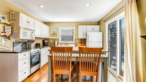 Cocina con armarios blancos, microondas, horno y refrigerador. Hay dos sillas de madera junto a una mesa, y una ventana con persianas.