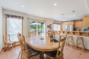 A dining area with a wooden table and chairs, a kitchen with wooden cabinets, and a view of the outdoors through large windows. - Sweet Sunriver Home with Private Hot Tub, Outdoor Space & Eight SHARC Passes (Sunriver)