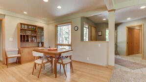 A dining area with a wooden table and chairs, a glass cabinet with kitchenware, and a large window with a view of trees.