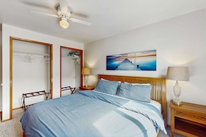 Une chambre à coucher comprenant un lit, un ventilateur de plafond, une table de chevet avec une lampe, une tête de lit en bois, une œuvre d’art accrochée au mur et une armoire avec un miroir.