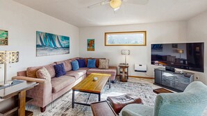 A living room with a sectional sofa, a coffee table, a TV, a ceiling fan, and artwork on the walls. - Delightful Home with Ocean View & Private Hot Tub - 3 Blocks to Beach (Yachats)