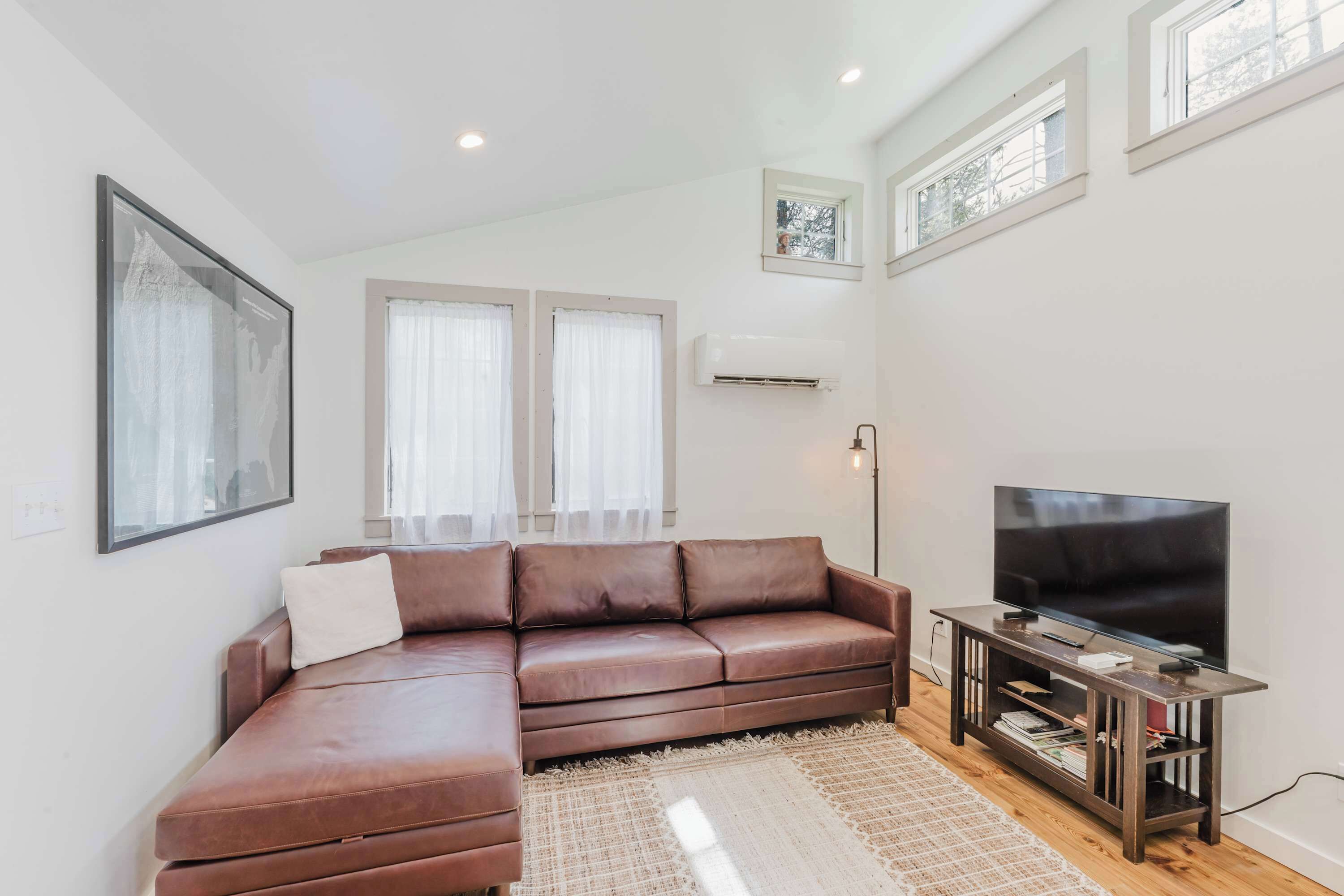 A modern living room with a brown leather sofa, a flat-screen TV on a wooden stand, and a large window with sheer curtains.