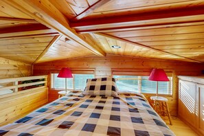 Un dormitorio de cabaña de madera con colcha a cuadros, dos lámparas rojas y una ventana con persianas.