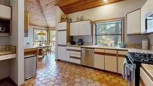 Cozinha com teto de madeira, piso de azulejos hexagonais, área de jantar com cadeiras e janela com vista.