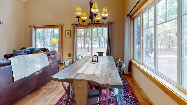 Un comedor con mesa de madera, sillas, un candelabro y ventanas amplias con vistas a árboles.