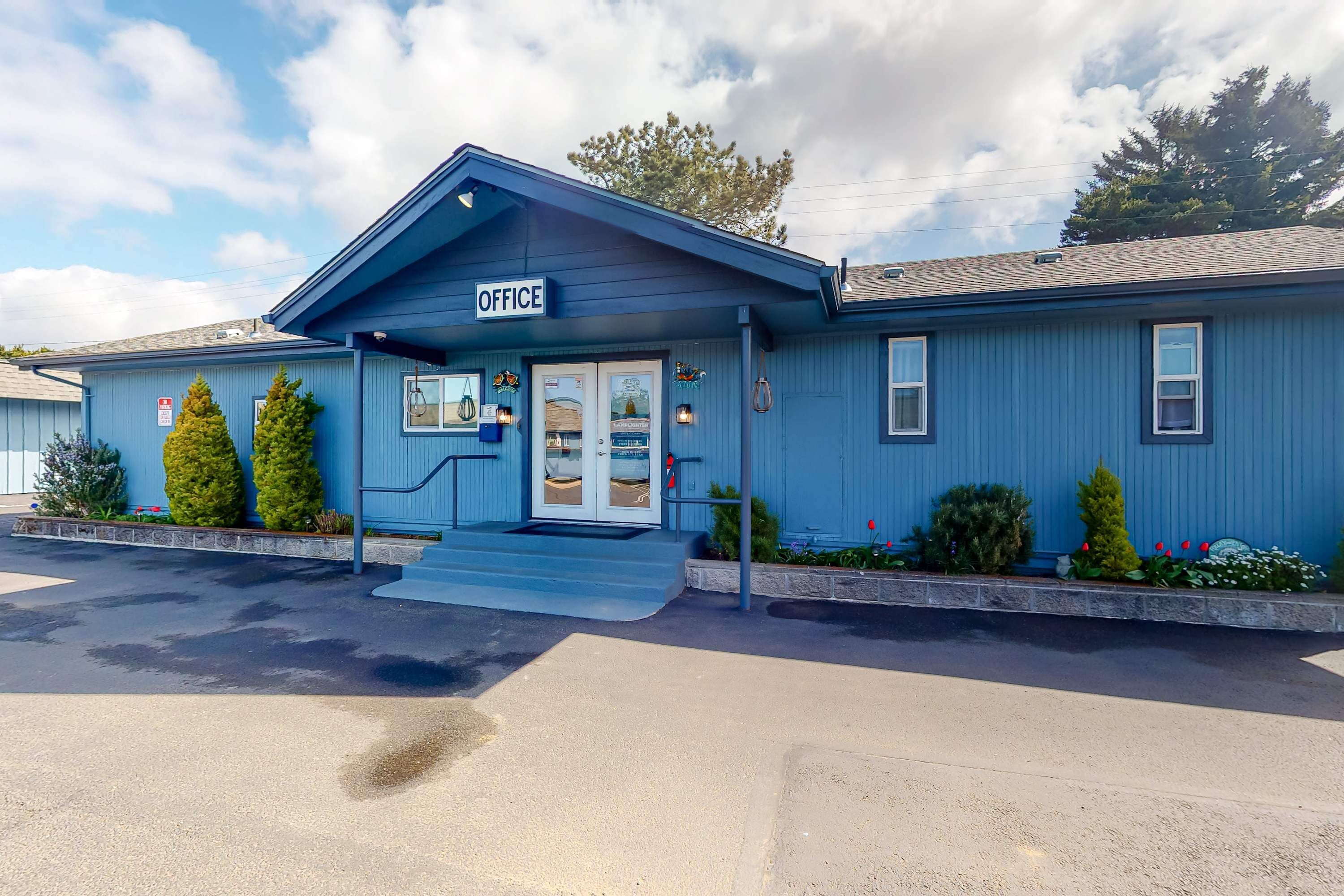 A blue building with a sign reading 'OFFICE' at the entrance, a set of steps leading up to the door, and a small porch area with potted plants.