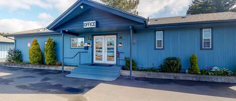A blue building with a sign reading 'OFFICE' at the entrance, a set of steps leading up to the door, and a small porch area with potted plants.