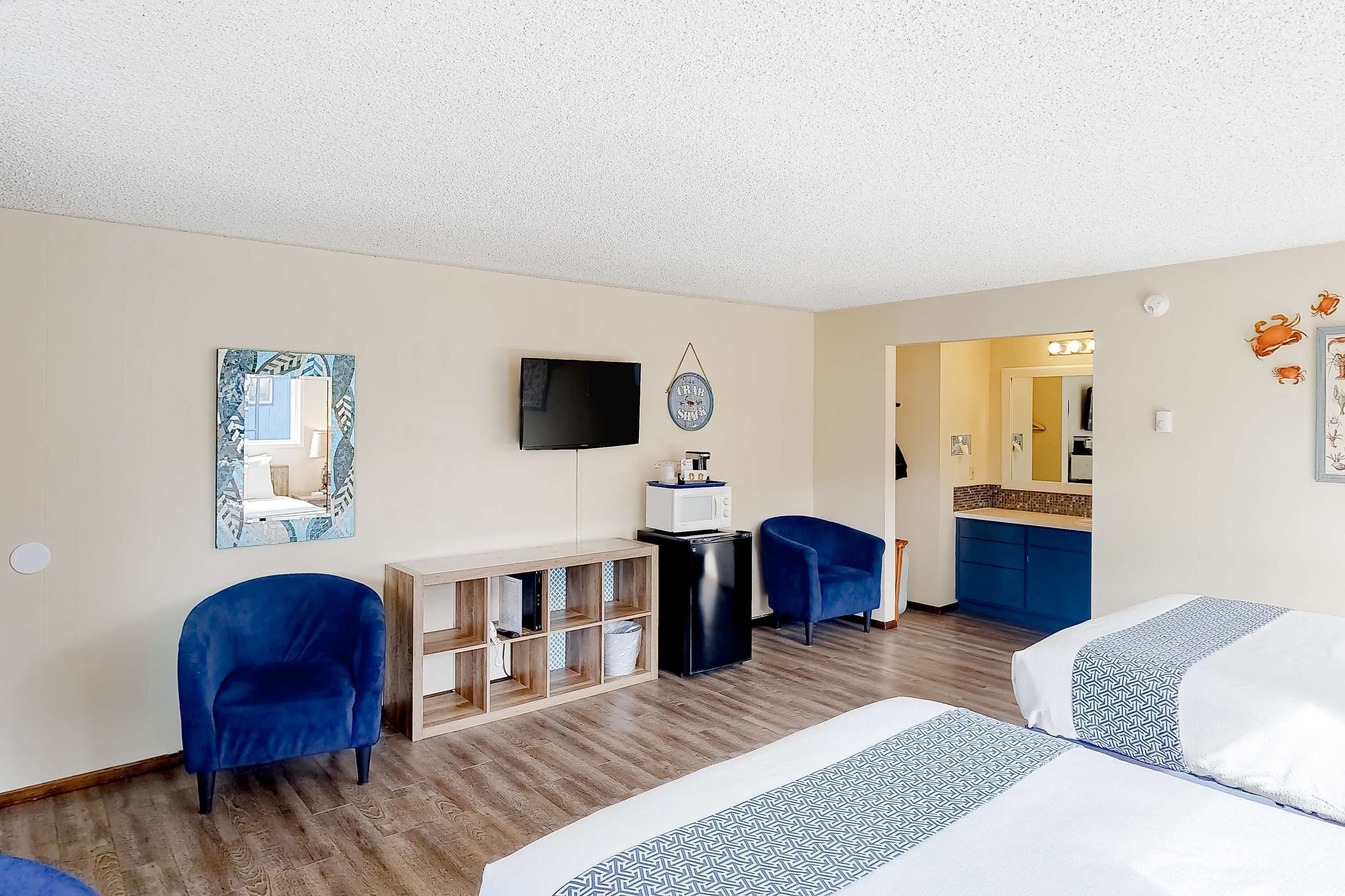 A hotel room with two beds, a TV, a small kitchenette, a mirror, and a blue chair.