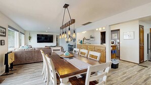 Een moderne eetruimte met een houten tafel en witte stoelen, een keuken met houten kastjes, en een groot raam dat natuurlijk licht binnenlaat.