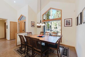 A dining area with a large wooden table, six chairs, a chandelier, and a view of trees through a large window. - Cozy home near SHARC has AC with a large deck and gas grill (Sunriver)