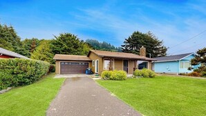A single-story house with a driveway leading to a garage, surrounded by well-maintained lawns and trees. - Experience Comfort and Serenity by the Sea, Dog Friendly with AC, & Patio (Yachats)