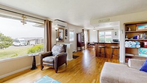 A spacious living area with a large window, a leather armchair, a sofa, a small round table, and a bookshelf. - Experience Comfort and Serenity by the Sea, Dog Friendly with AC, & Patio (Yachats)
