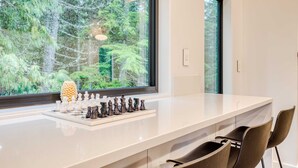 A modern kitchen with a chessboard on the counter, large windows with a forest view, and two chairs.