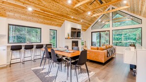 A modern interior with a wooden ceiling, large windows, a dining area, and a living space with a sofa. - Sleek, Modern Home in Town with Forest Views, Fire Table, WiFi - Walk to Skiing (Government Camp)