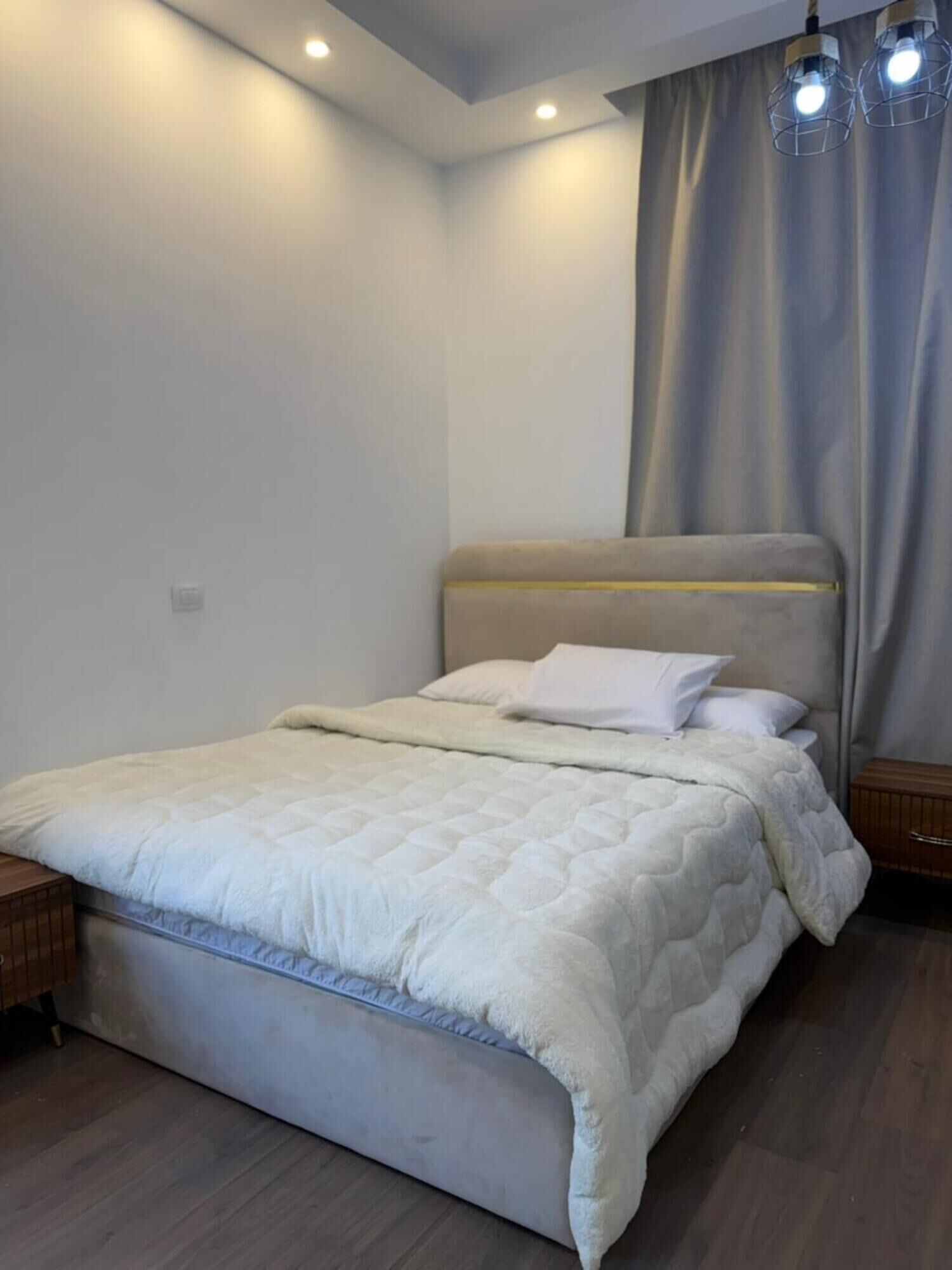 Un dormitorio con cama, cabecera, mesita de noche, suelo de madera y cortina.