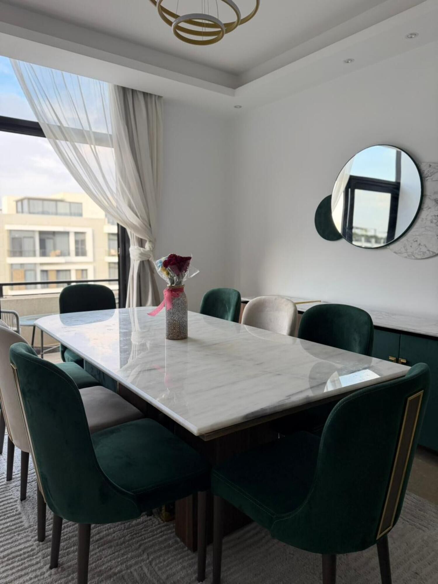 A dining area with a marble table, green upholstered chairs, a vase with flowers, a round mirror, and a view of buildings through sheer curtains.