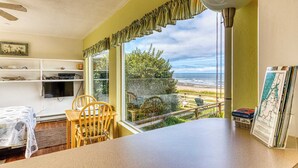 A room with a view of the beach, a dining area with a table and chairs, a shelf with items, and a window with curtains.