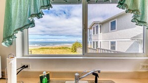 A kitchen with a window view of a beach and a house, a paper towel holder, a soap dispenser, and a faucet.