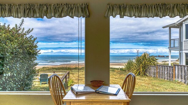 A room with a table and chairs facing a beach view through large windows.