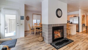 A living room with a fireplace, wooden flooring, a dining area with chairs, and a kitchen with a refrigerator. - Golf course-view townhome with private hot tub & SHARC access (Sunriver)
