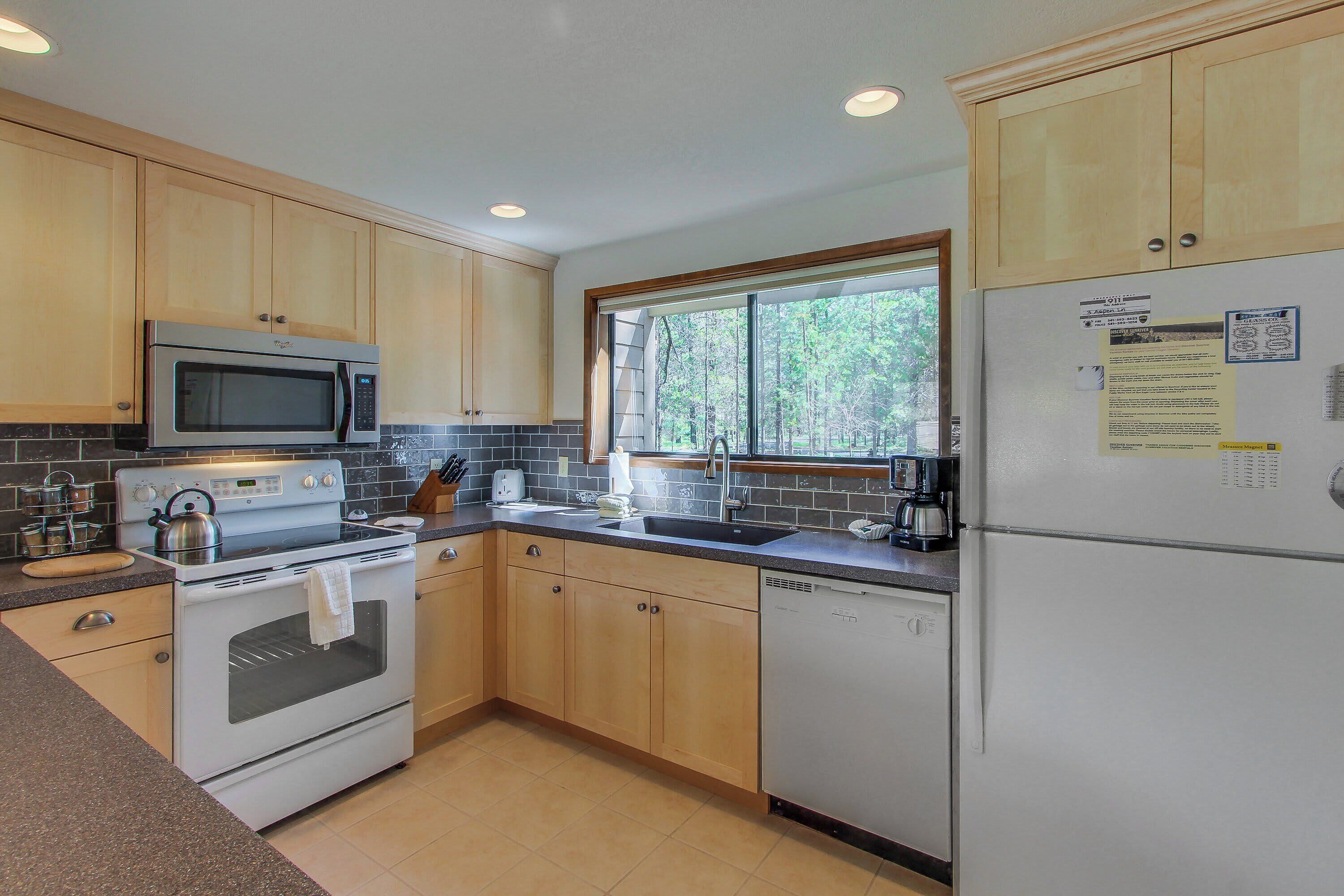 A kitchen with wooden cabinets, a stainless steel microwave, an oven, a dishwasher, and a window with a view of trees.