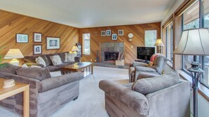 A living room with a fireplace, wooden walls, and a large window. - Cabin with Private Hot Tub Near the Deschutes River - Free SHARC access (Sunriver)