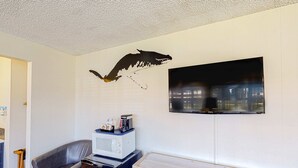 A room with a mounted TV, a microwave on a cabinet, a grey sofa, and a wooden table. - Retro beach suite with  fast WiFI - walkable to downtown hot spots (Bandon)