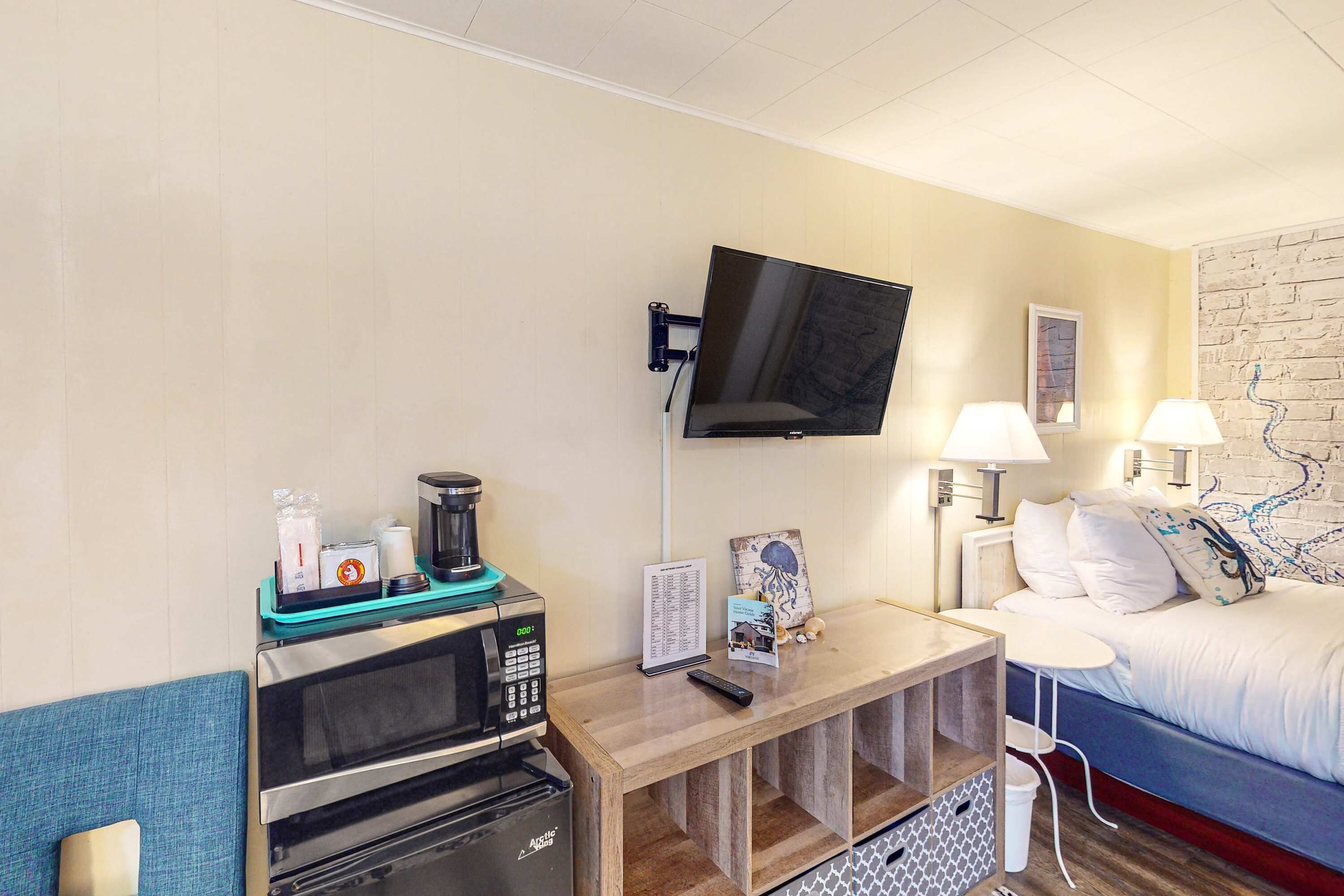 A hotel room with a flat-screen TV mounted on the wall, a microwave, a coffee maker, a small table with a remote control, a bed with white bedding, and a nightstand with a lamp.