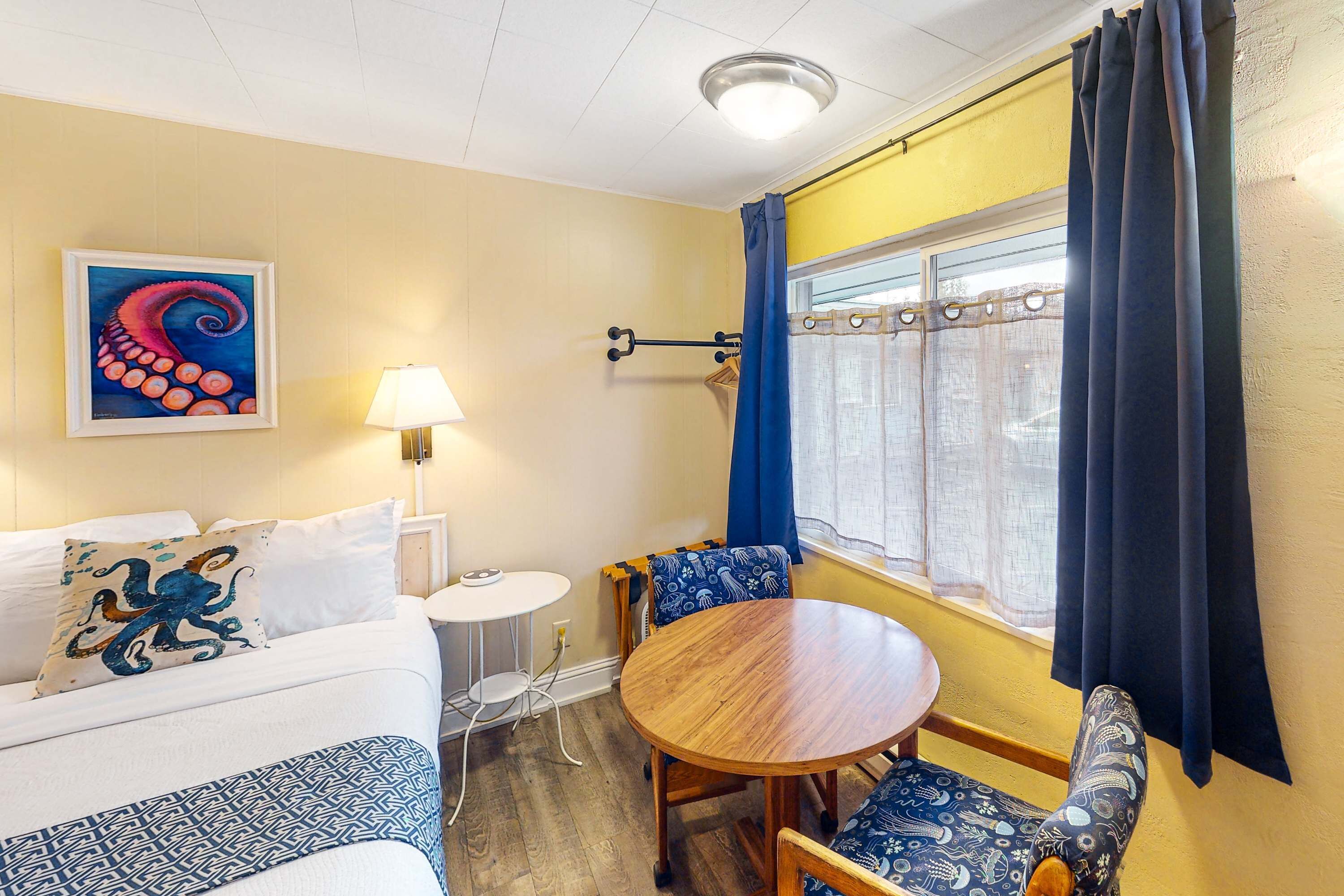 A room with a bed, a round wooden table, blue patterned chairs, a lamp, a painting, and a window with curtains.