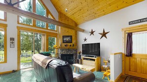 A spacious living room with a fireplace, a television, and a large window overlooking a forest. - Inviting chalet with private hot tub, grill, 3 pools, hot tub, gym, & tennis (Eagle Crest)