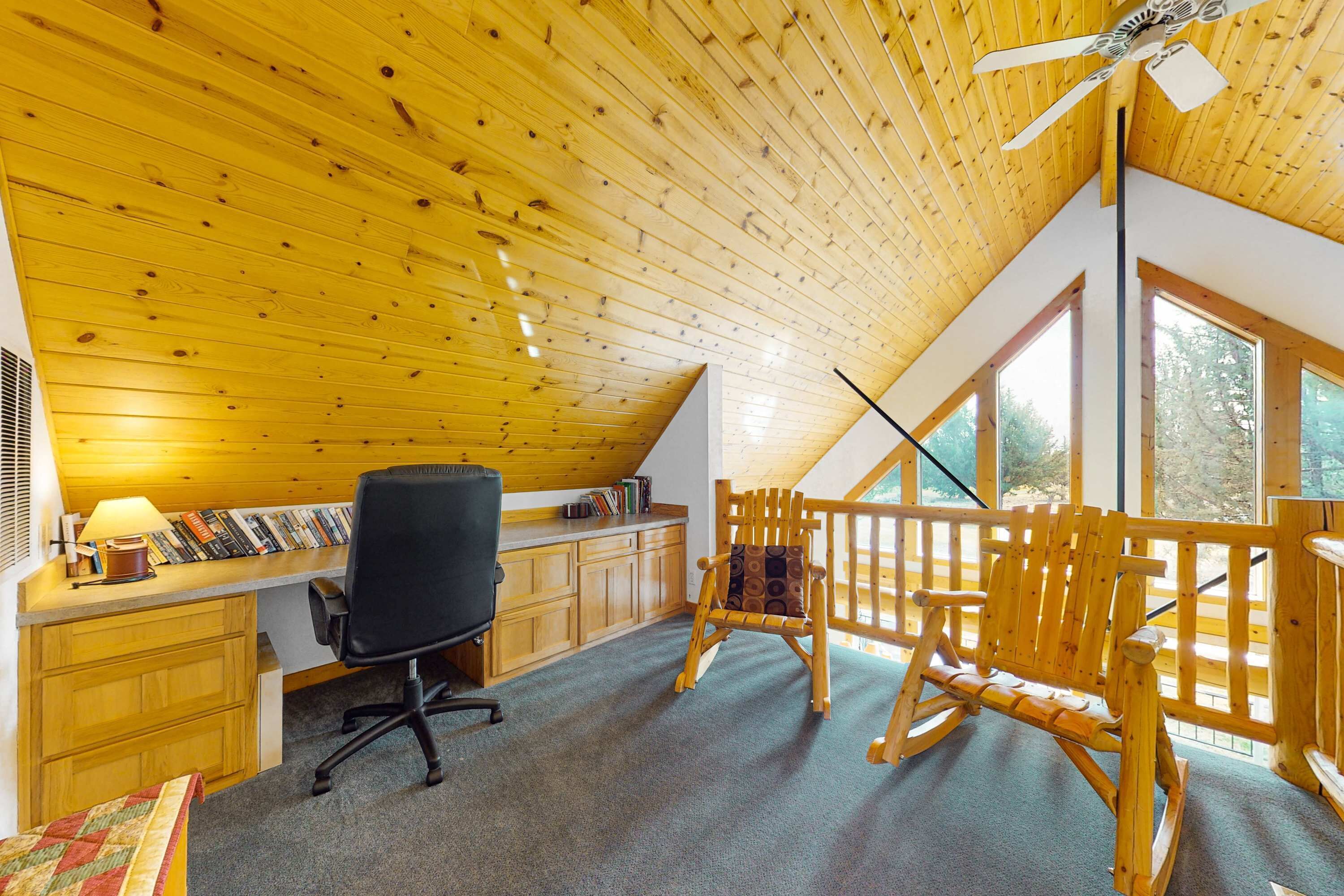 A cozy room with a sloped ceiling, wooden desk, office chair, rocking chair, and large windows.
