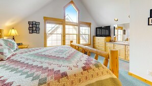 A bedroom with a large bed, a wooden headboard, a window with a view, a television, and a kitchen area with wooden cabinets. - Inviting chalet with private hot tub, grill, 3 pools, hot tub, gym, & tennis (Eagle Crest)