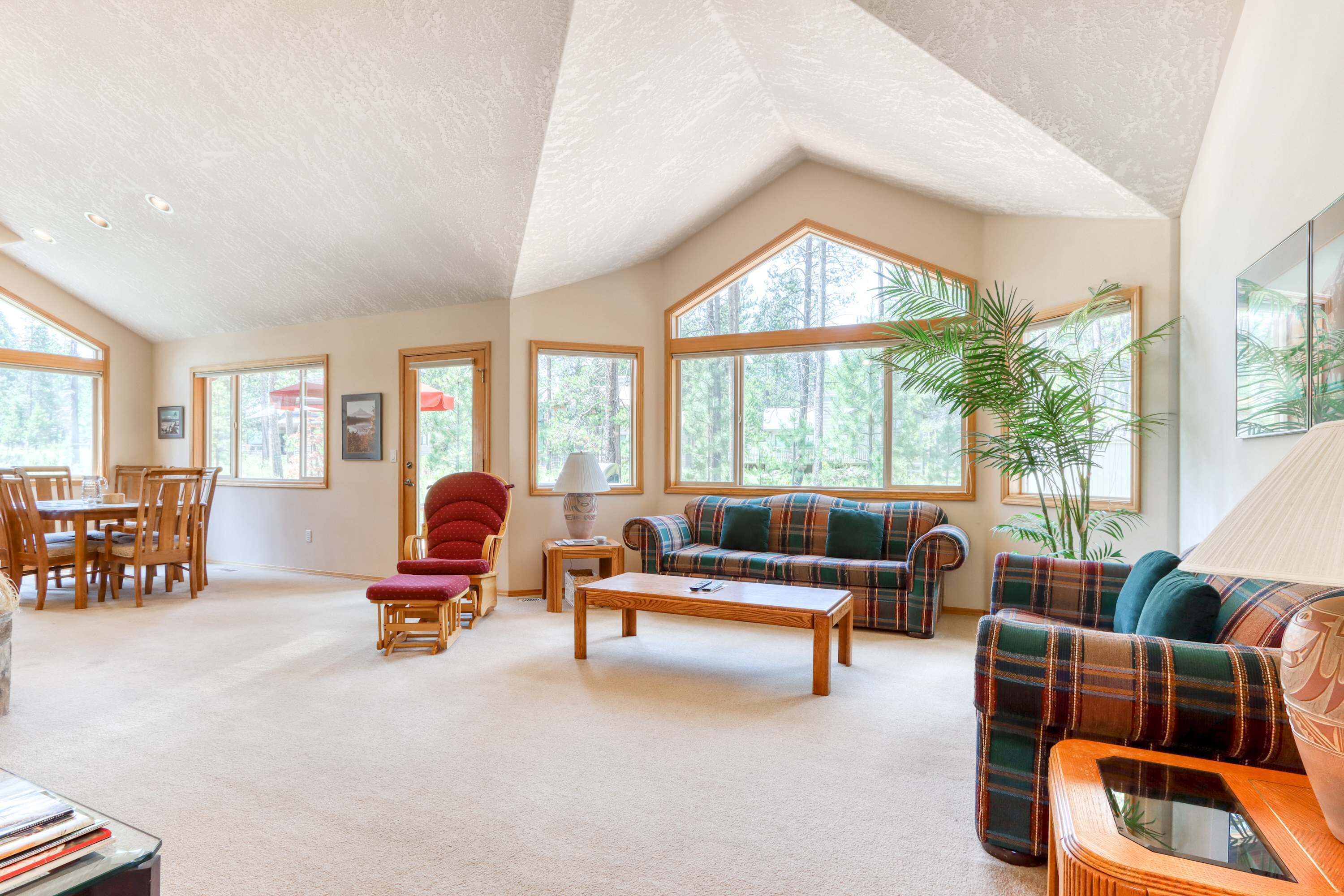 A spacious living room with a high ceiling, large windows, a plaid sofa, a red chair, and a dining area with wooden furniture.
