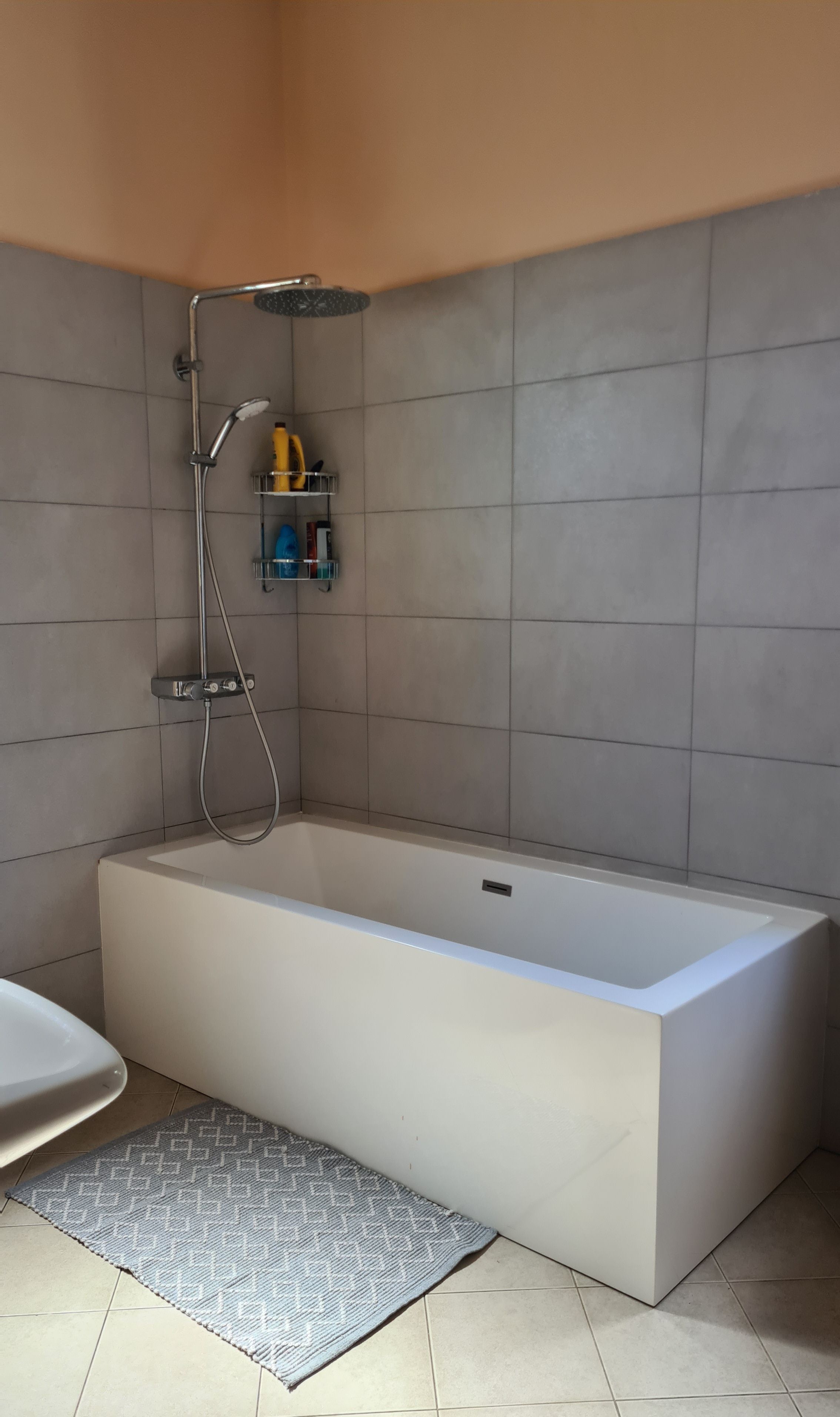 A modern bathroom with a large rectangular bathtub, a showerhead, and a small shelf with toiletries.
