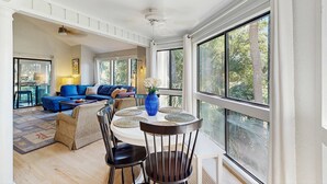 A spacious living area with a blue sofa, a round table with chairs, and large windows offering a view of trees. - Home with patio, golf views, close to beach and restaurants (Edisto Beach)