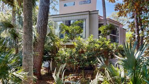 A modern house with a flat roof, large windows, and surrounded by various trees and plants. - Home with patio, golf views, close to beach and restaurants (Edisto Beach)