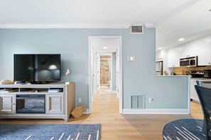 A modern living room with a flat-screen TV, a fireplace, and a kitchen area with white cabinets and appliances. - Harbourtown Gem (Hilton Head)
