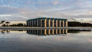 Un hôtel à plusieurs étages, doté d’une piscine dont la surface réfléchit la lumière, situé près d’une plage.