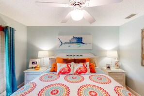 A bedroom with a bed, two bedside tables, a ceiling fan, a wall-mounted artwork, and a window with curtains. - Just Trippin' (Edisto Beach)