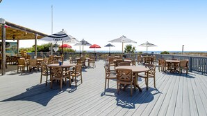 Eine Terrasse im Freien mit Holztischen und -stühlen, Sonnenschirmen und Blick aufs Meer.