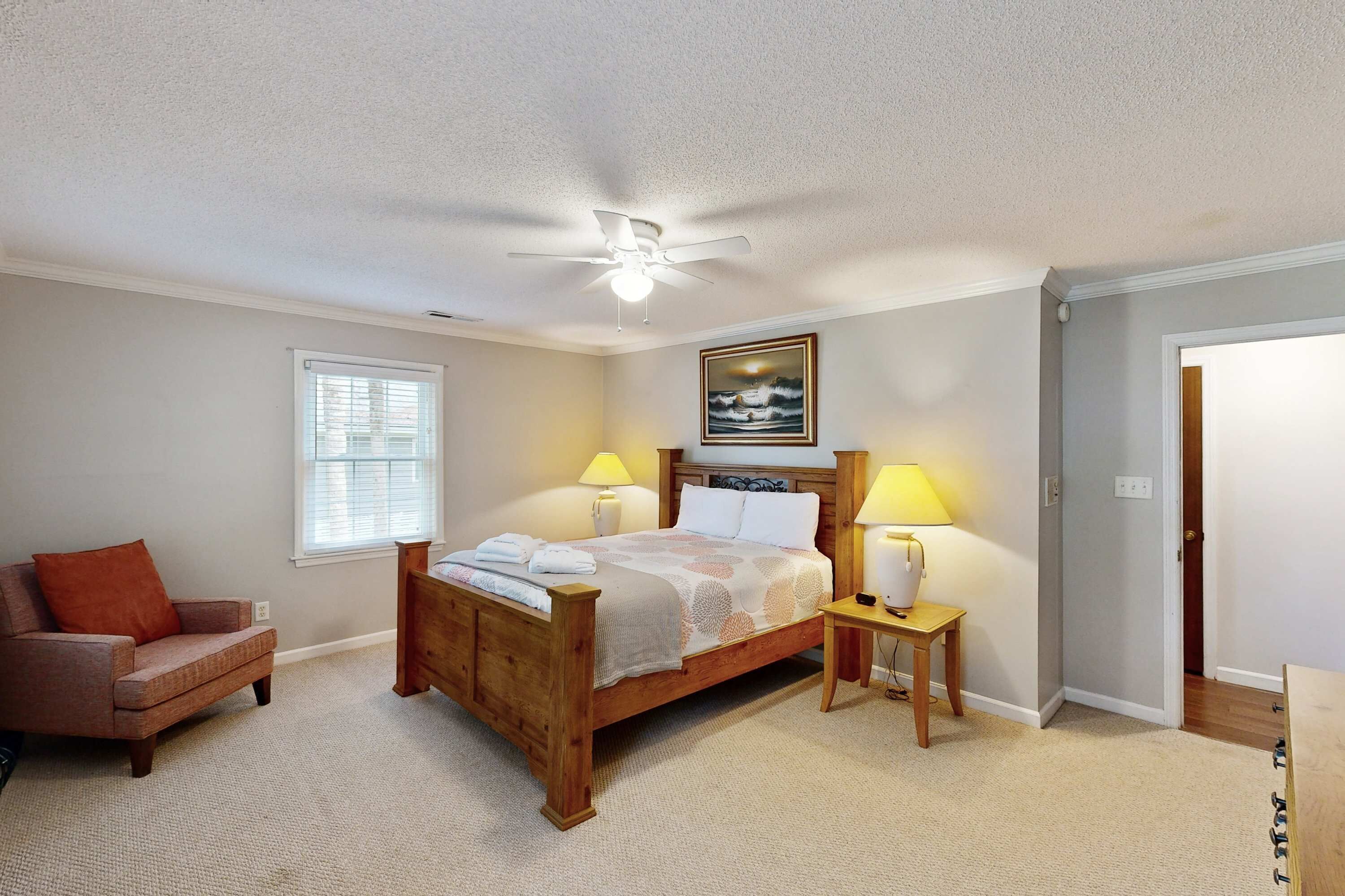 A bedroom with a wooden bed, two bedside tables with lamps, a chair, a ceiling fan, and a window with blinds.