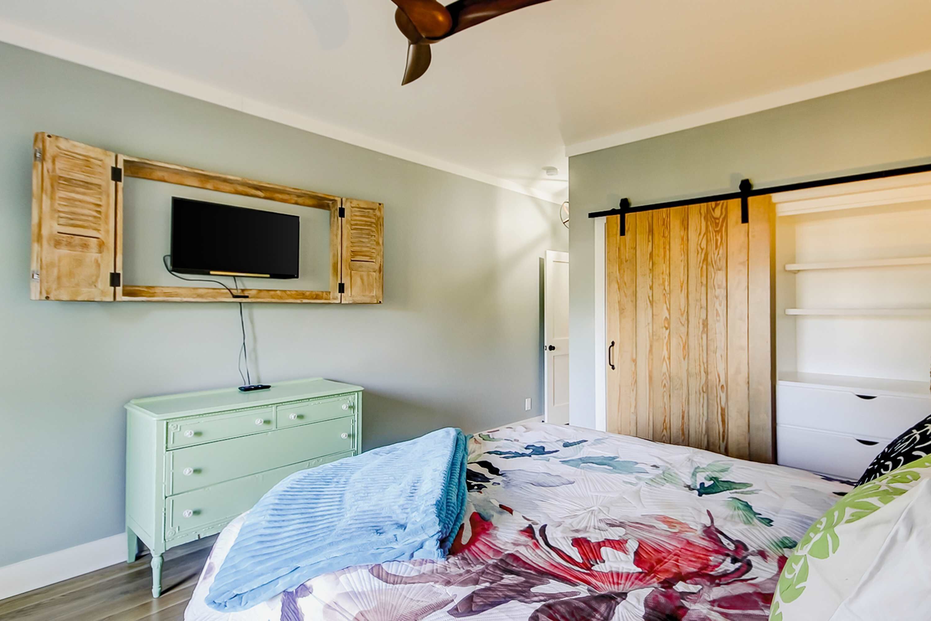 Une chambre à coucher comprenant un lit, un téléviseur fixé sur un cadre en bois, une commode et une armoire à porte coulissante.