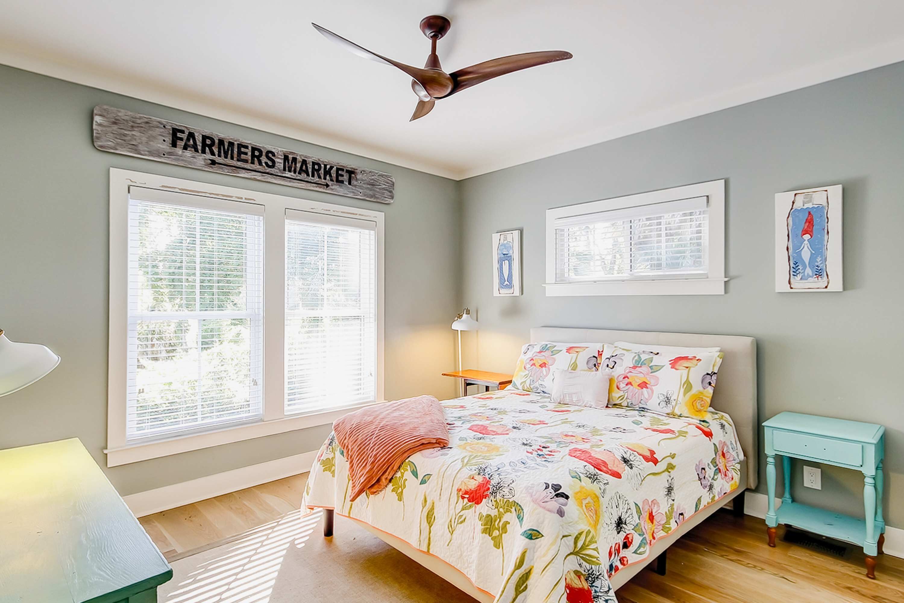 A bedroom with a bed, a ceiling fan, a window with blinds, a bedside table, and a wall sign.