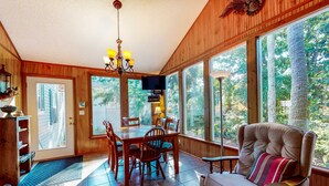 A dining area with a wooden table and chairs, a cushioned armchair, and large windows offering a view of trees. - Beautiful home within walking distance to the Beach w/ Free WiFi, Sun Deck (Edisto Beach)