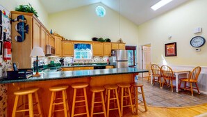 A kitchen with wooden cabinets, a central island with stools, a dining area with chairs, and a clock on the wall. - Beautiful home within walking distance to the Beach w/ Free WiFi, Sun Deck (Edisto Beach)