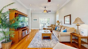 A spacious living room with wooden flooring, a large flat-screen TV, a ceiling fan, a sofa, a coffee table, and a large potted plant.